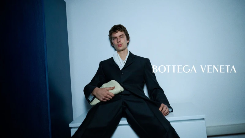 Leon Dame in a black tailored suit seated with a woven clutch bag in a Bottega Veneta campaign