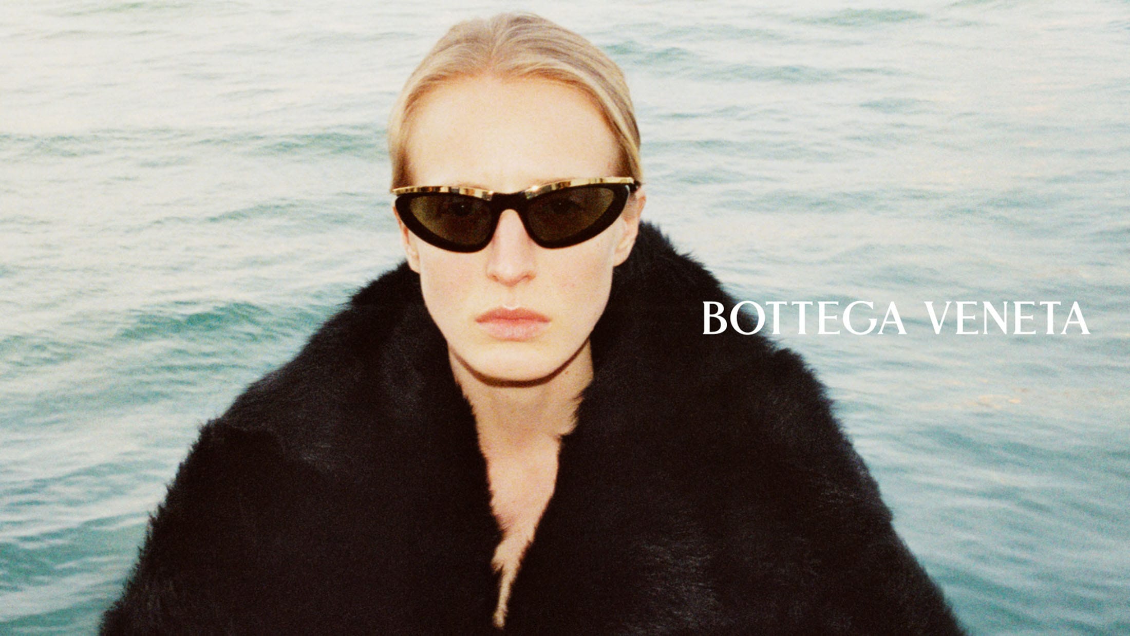 Model wearing black sunglasses with gold accents and a fuzzy black coat against open water for Bottega Veneta.
