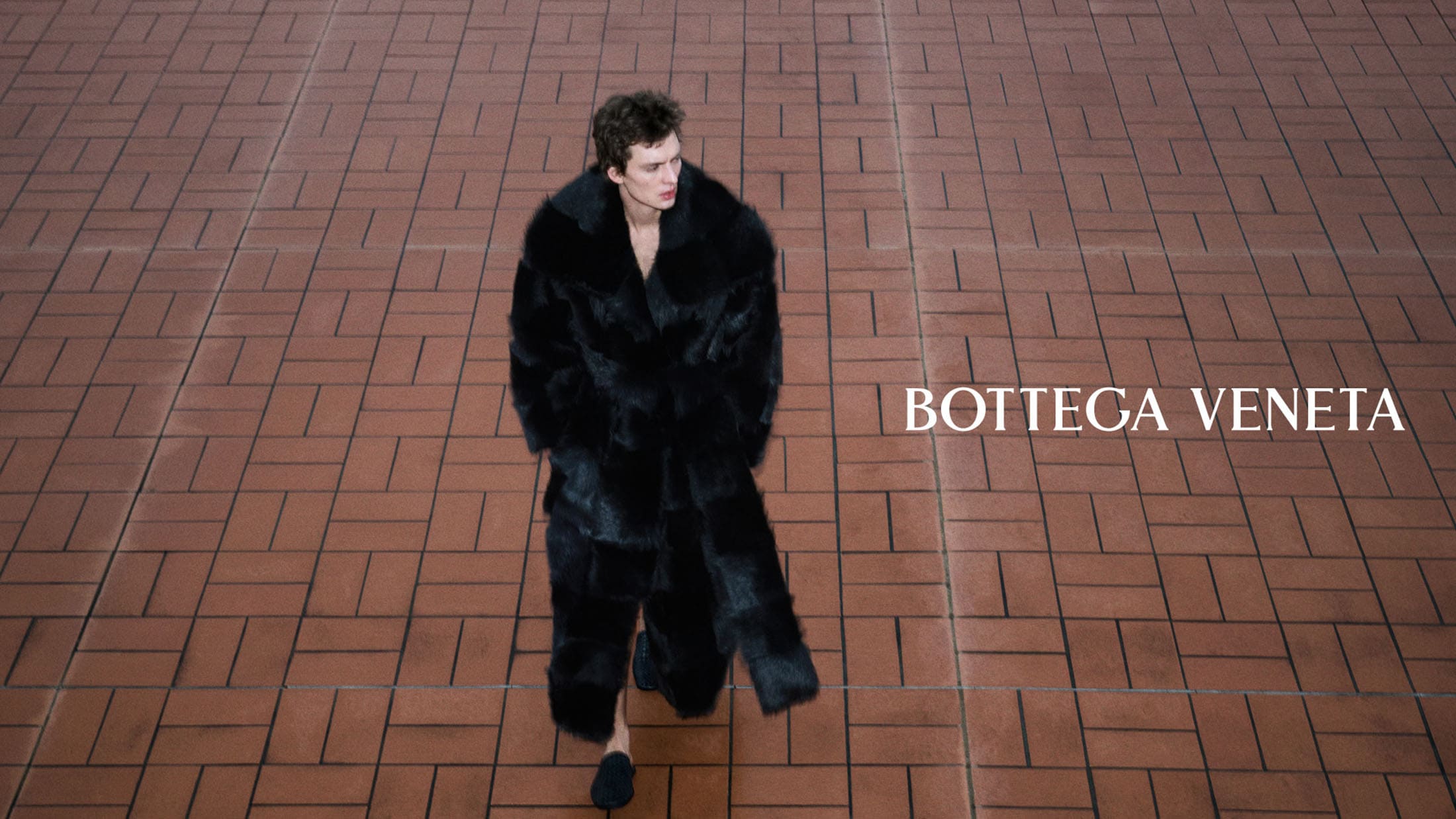 High-angle shot of a model walking across red brick pavers wearing an oversized black fur coat for Bottega Veneta.