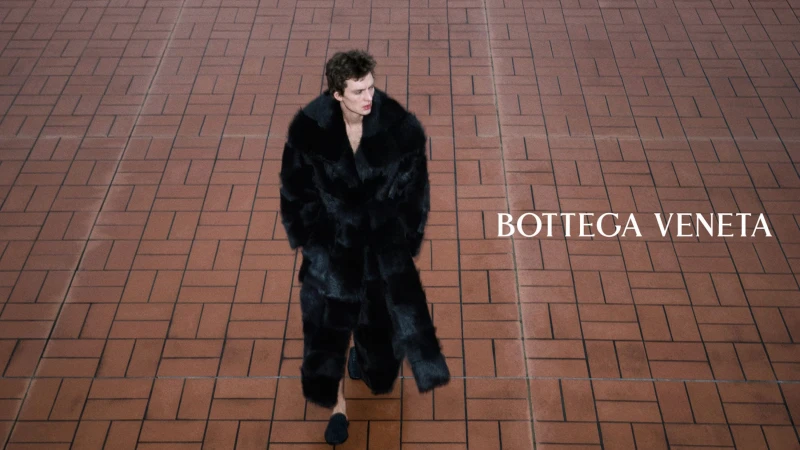 High-angle shot of a model walking across red brick pavers wearing an oversized black fur coat for Bottega Veneta.