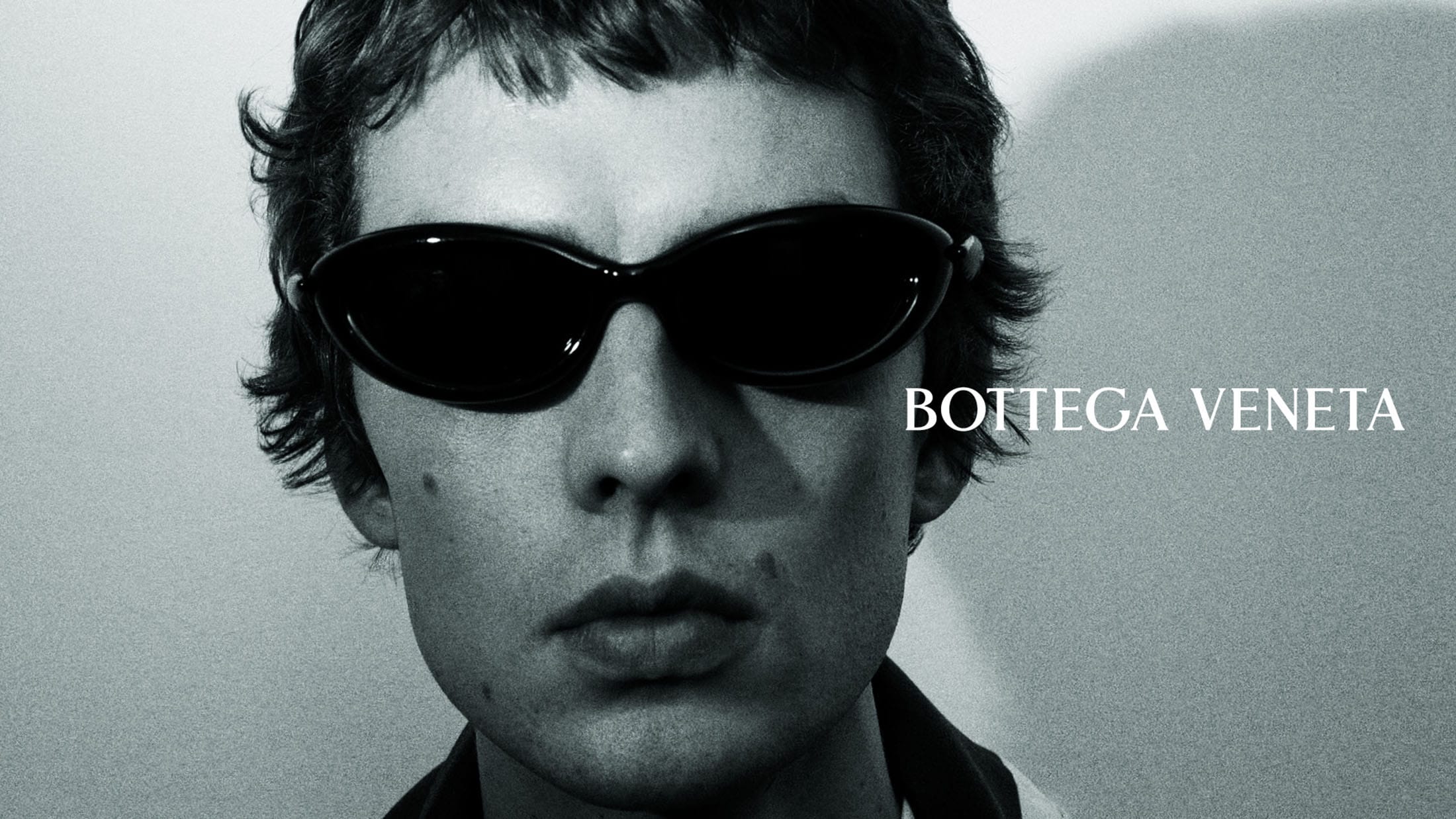 Close-up portrait of model Leon Dame wearing black oval sunglasses against a neutral studio backdrop.