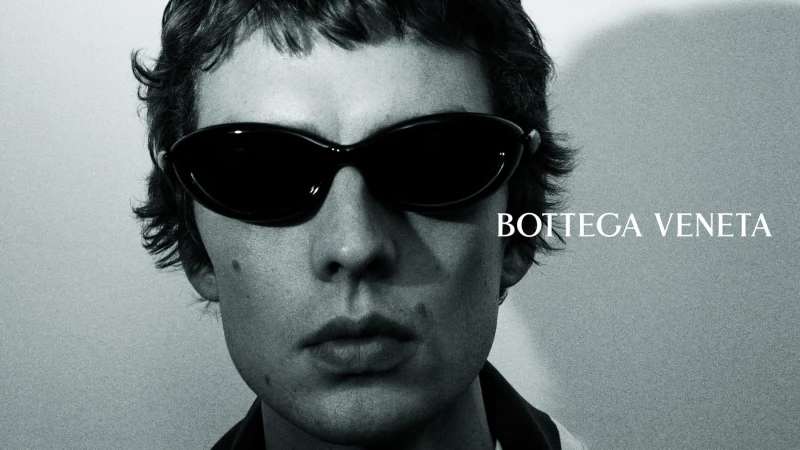 Close-up portrait of model Leon Dame wearing black oval sunglasses against a neutral studio backdrop.