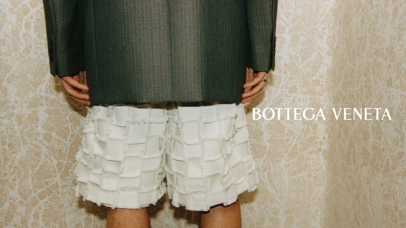 Model seen from behind wearing a charcoal pinstripe blazer and white textured fringed shorts against a marbled wall.