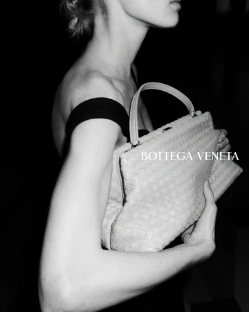Close-up black and white photo of a woman holding a pale woven leather Bottega Veneta handbag against a dark backdrop.