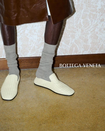 Close-up of legs wearing grey ribbed socks and cream woven leather slip-on loafers against a textured wall.