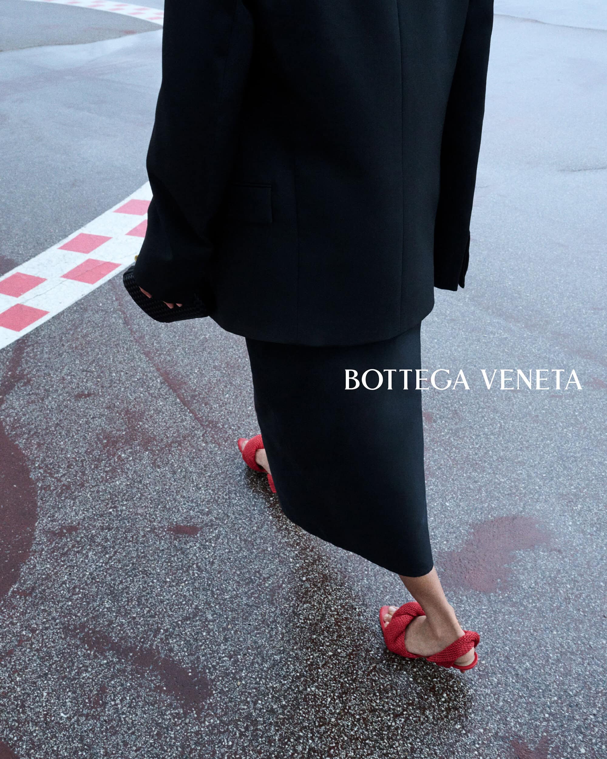 Rear low-angle view of a person walking across wet asphalt in a tailored black suit jacket, black midi skirt, and bright red slide sandals.