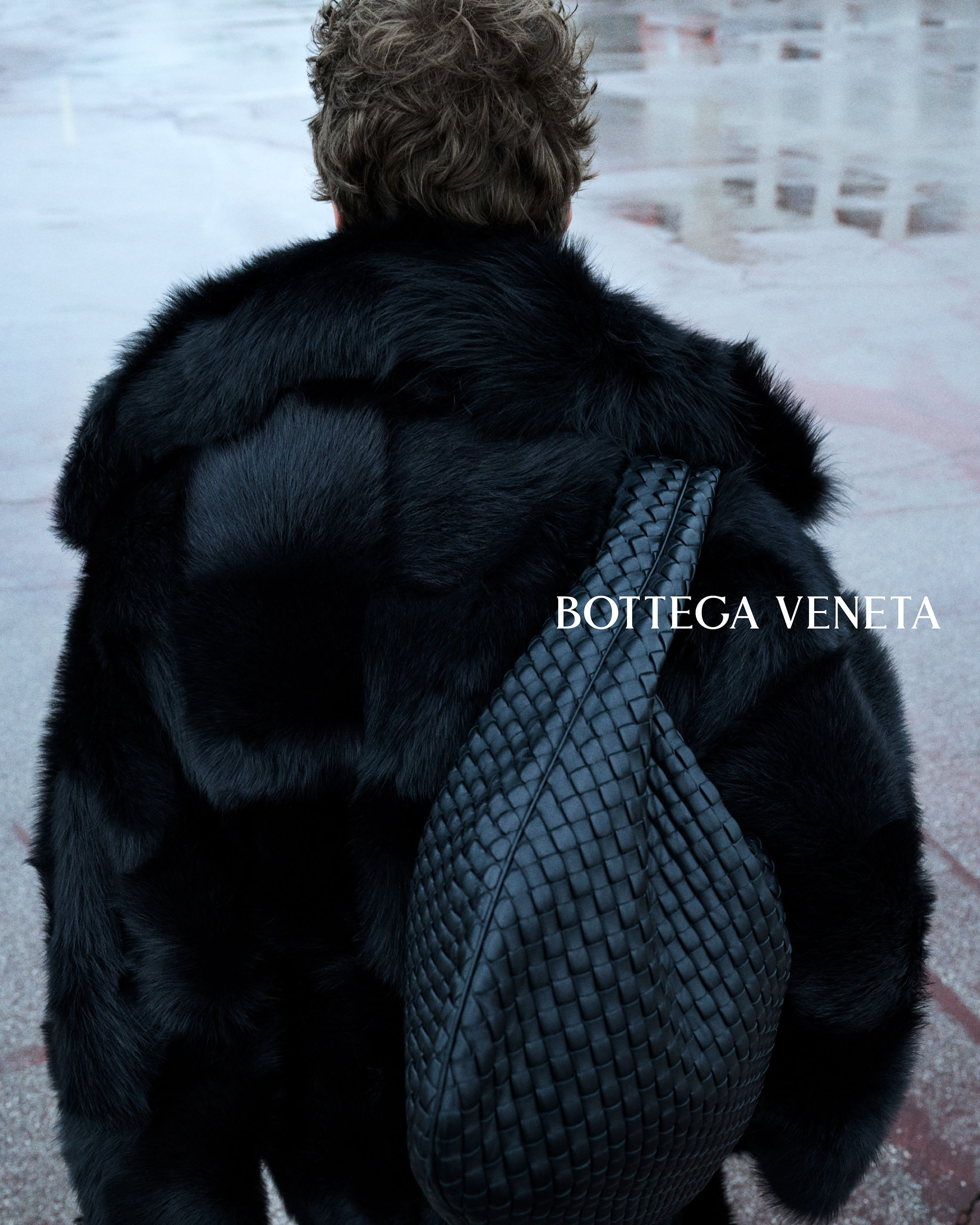 Model seen from behind wearing a black plush fur coat and an oversized black woven leather shoulder bag on wet pavement.