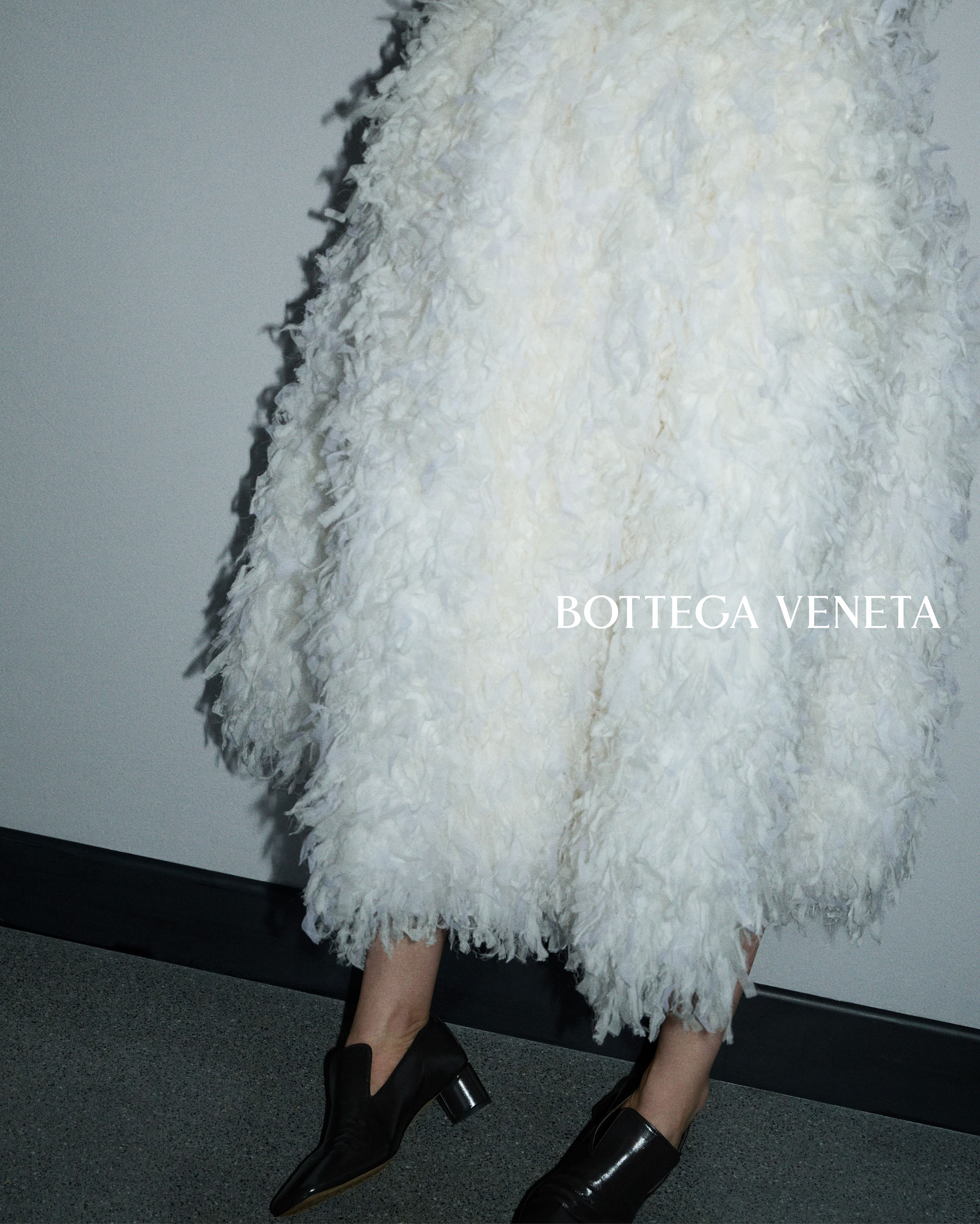 Model wearing a voluminous white feathered fringe dress and black leather block-heel pumps against a neutral wall.