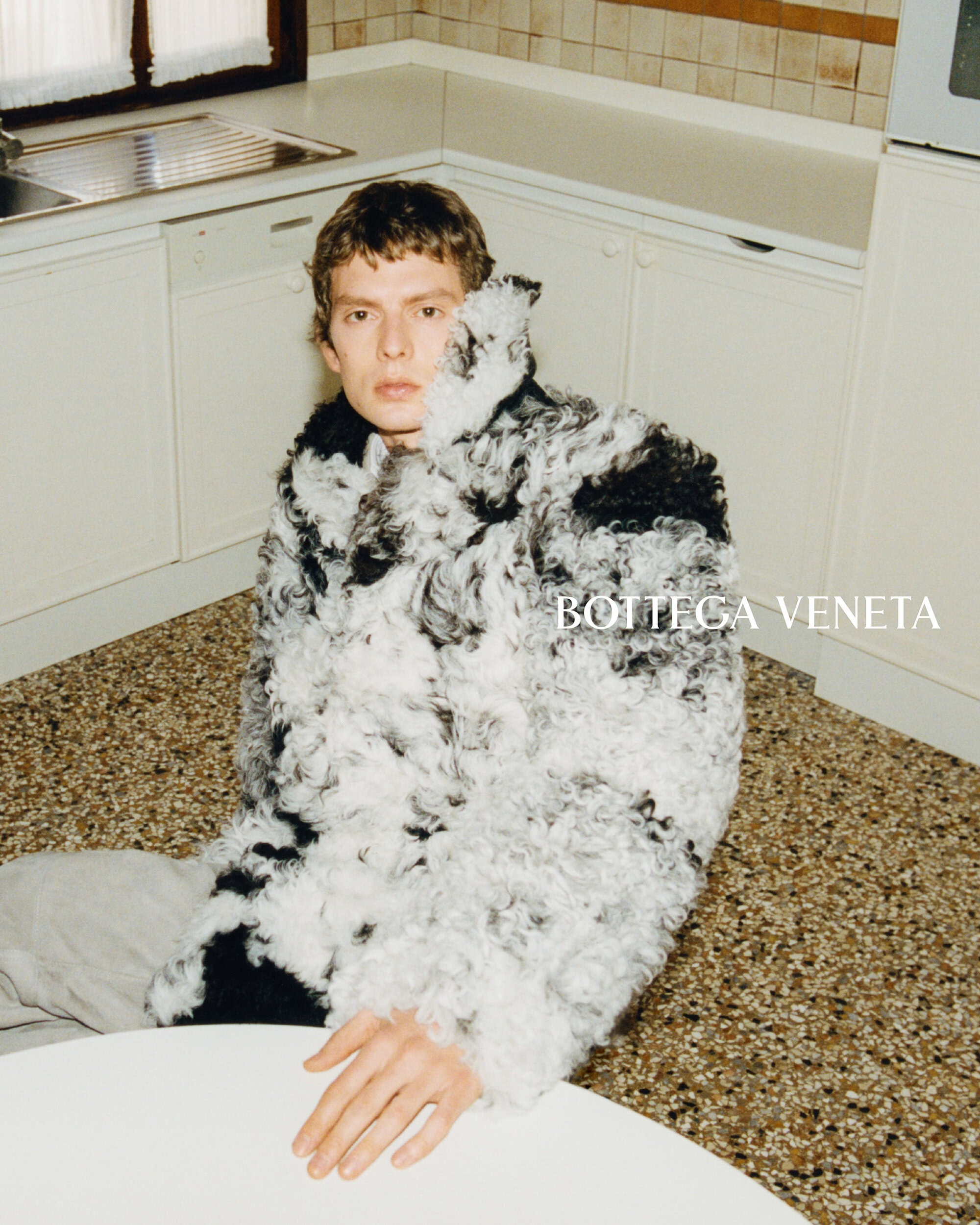 Man wearing a textured black and white coat seated in a retro kitchen setting with terrazzo flooring.