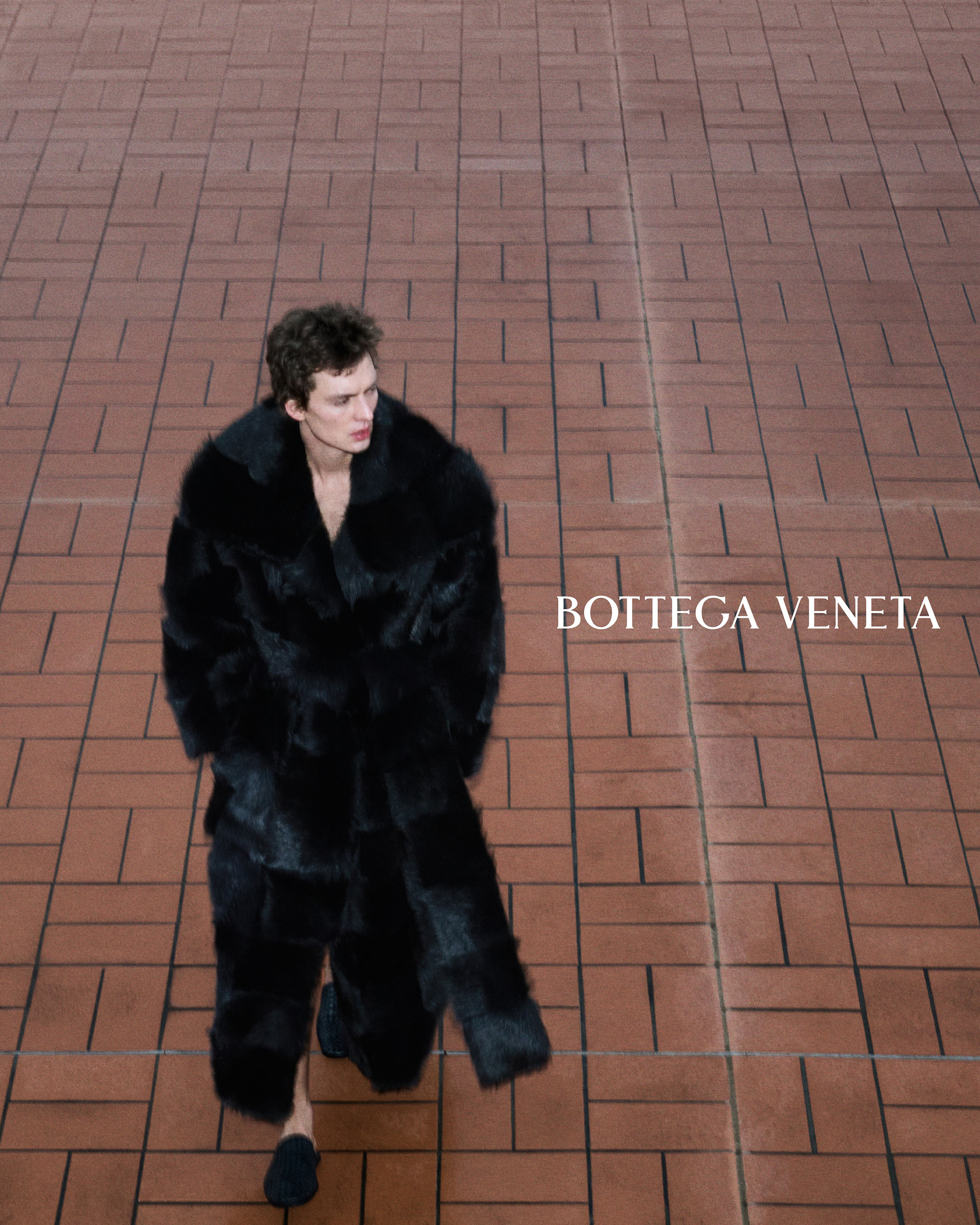 Model Leon Dame walking in a long black textured fur coat across a terracotta tiled surface.