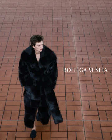 Model Leon Dame walking in a long black textured fur coat across a terracotta tiled surface.