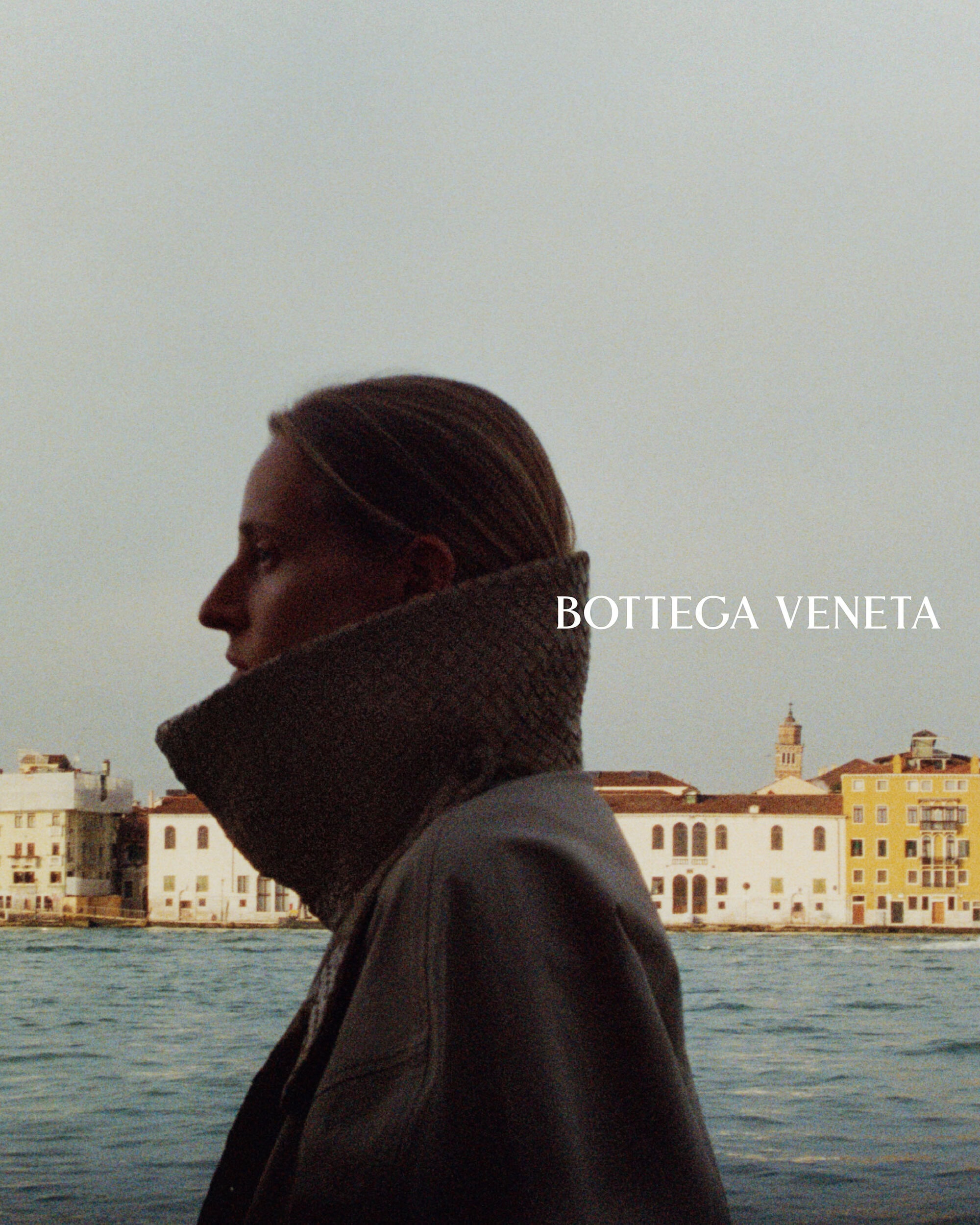 Profile view of a model wearing a high-collared coat against a waterfront cityscape backdrop.