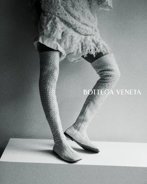 Model wearing textured fringe dress and woven thigh-high leather boots standing on a white plinth in a Bottega Veneta campaign.