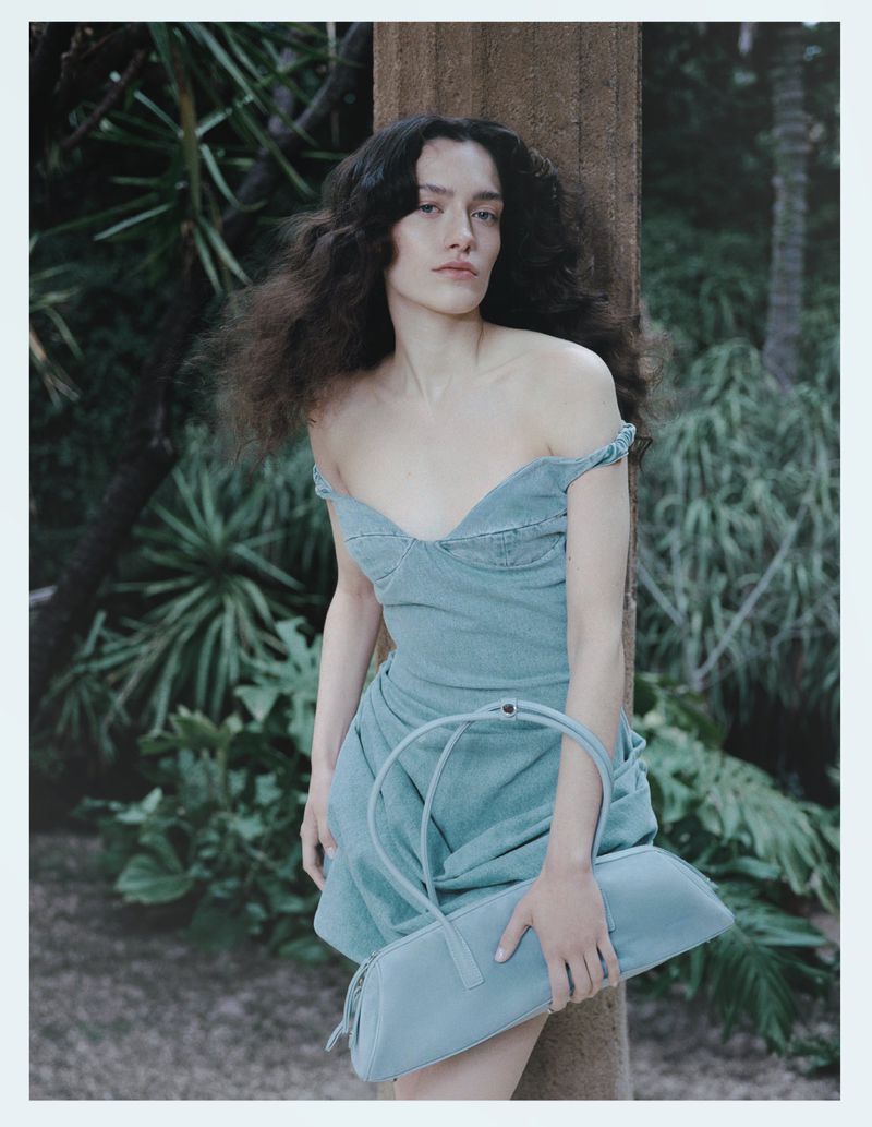 Model Julia Kuźniar in a light blue off-shoulder denim dress holding a matching elongated handbag against a pillar in a garden.