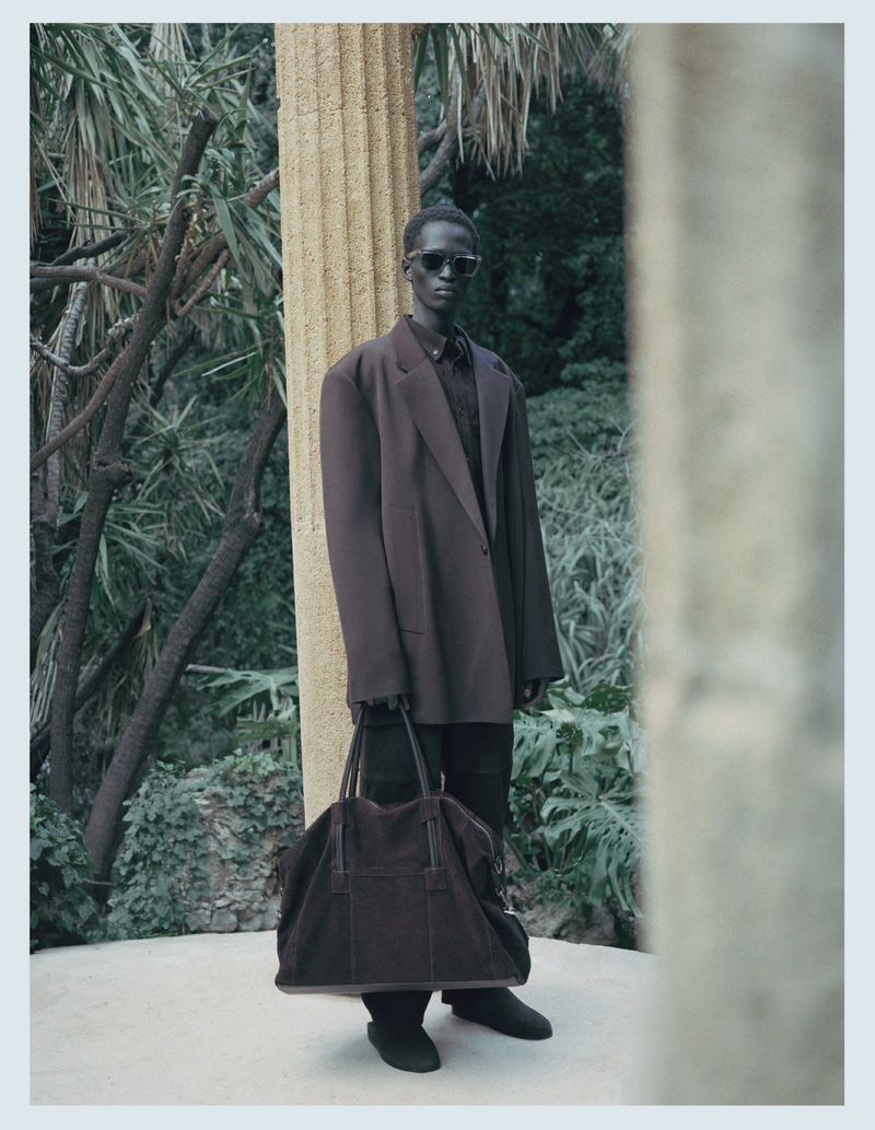 Man wearing a brown oversized blazer and sunglasses holding a large dark duffel bag outdoors among trees.