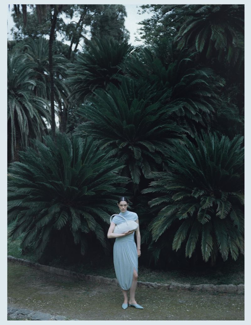Model Julia Kuźniar in a light blue draped dress holding an off-white shoulder bag in front of lush dark green palm trees.