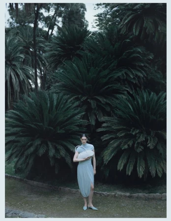Model Julia Kuźniar in a light blue draped dress holding an off-white shoulder bag in front of lush dark green palm trees.