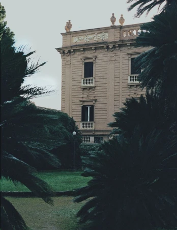 Classical villa facade with balconies viewed through dark green palm trees in an estate garden.