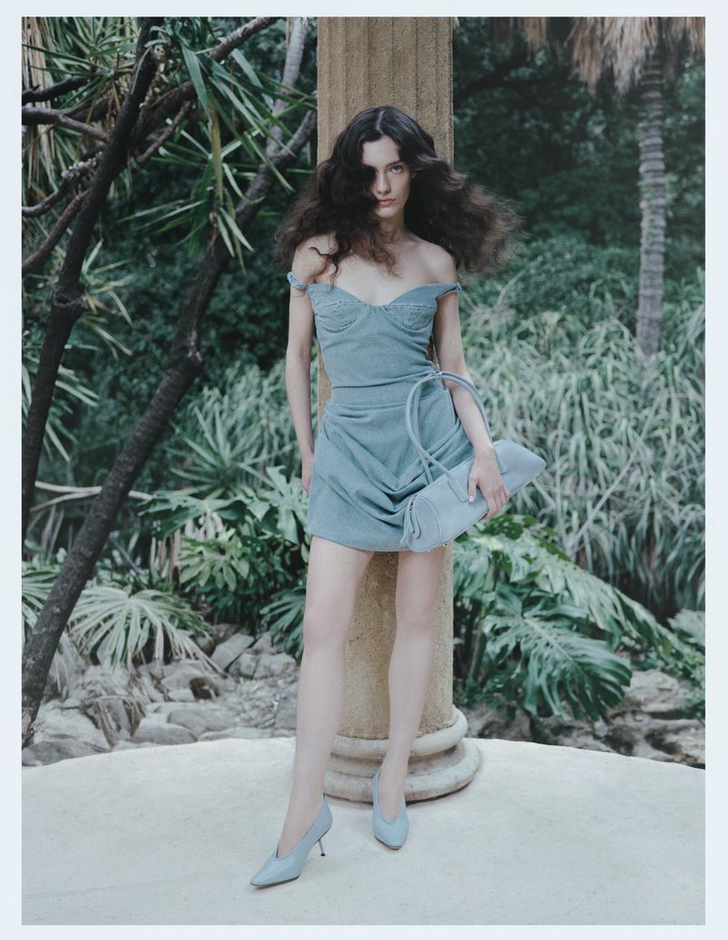 Model Julia Kuźniar wearing a light blue mini dress, pointed pumps, and shoulder bag leaning on a stone column outdoors.