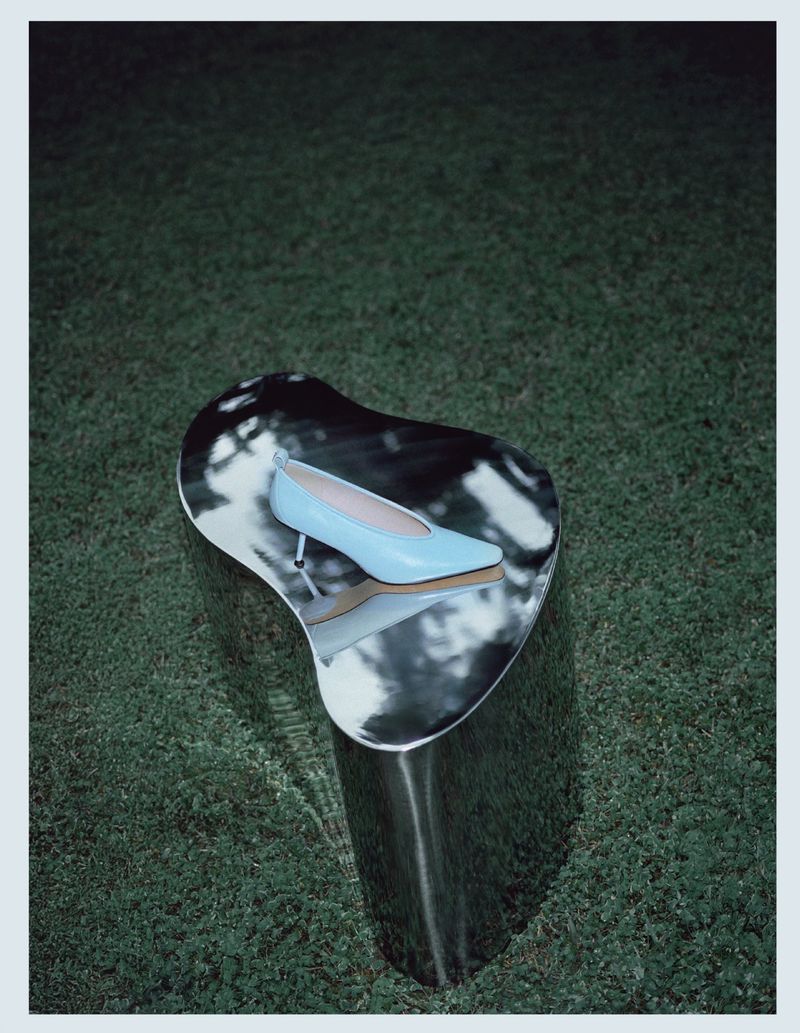 Light blue pointed kitten heel pump resting on a mirrored chrome pedestal set outdoors on green grass.