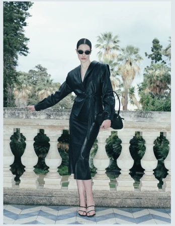 Model Julia Kuźniar wearing a black leather suit, dark sunglasses, and strappy heels against a stone balustrade with palm trees.