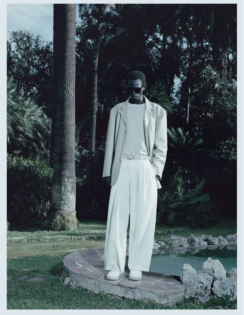 Model Mamuor Majeng wearing a relaxed cream suit and sunglasses while standing by a stone pool edge in a lush palm garden.