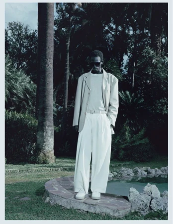 Model Mamuor Majeng wearing a relaxed cream suit and sunglasses while standing by a stone pool edge in a lush palm garden.