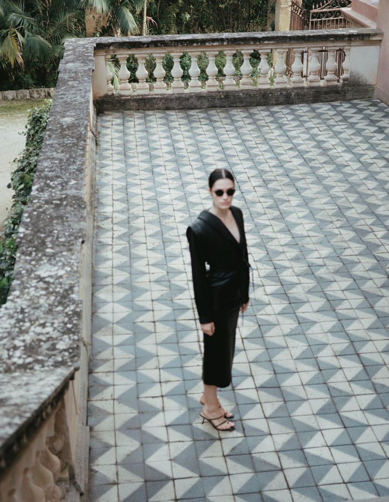 Model Julia Kuźniar in a black midi dress and sunglasses standing on a geometric tiled terrace beside a stone balustrade.