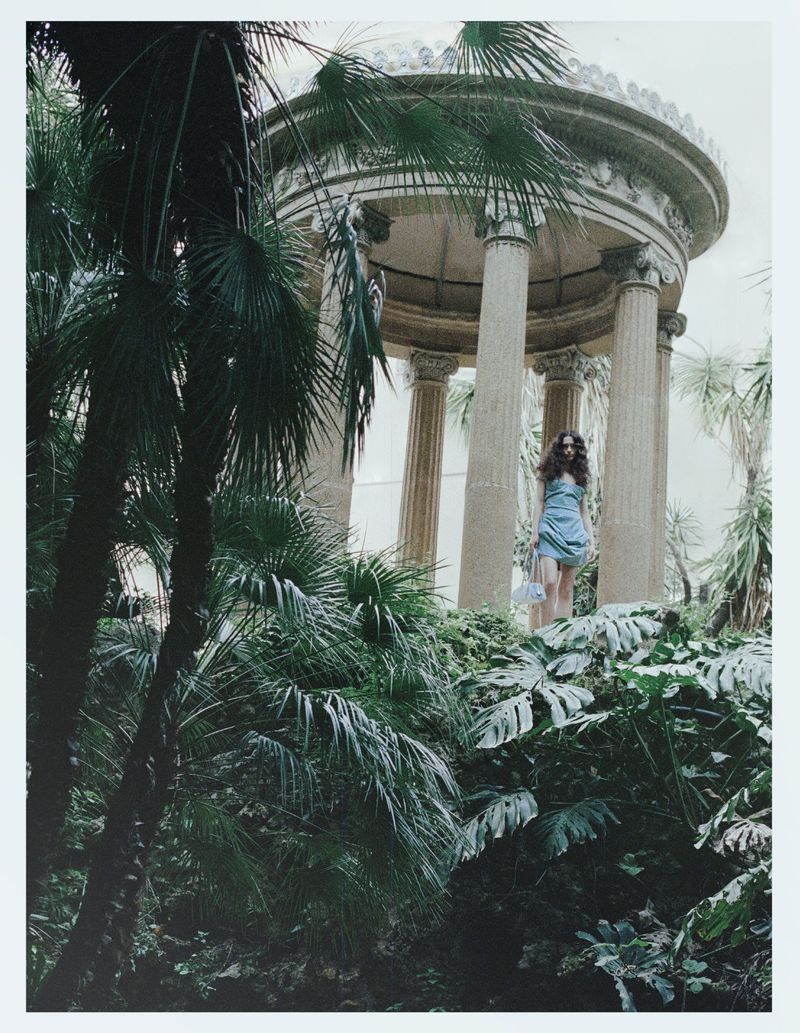 Model Julia Kuzniar standing in a powder blue mini dress by a classical stone rotunda framed by lush tropical foliage.