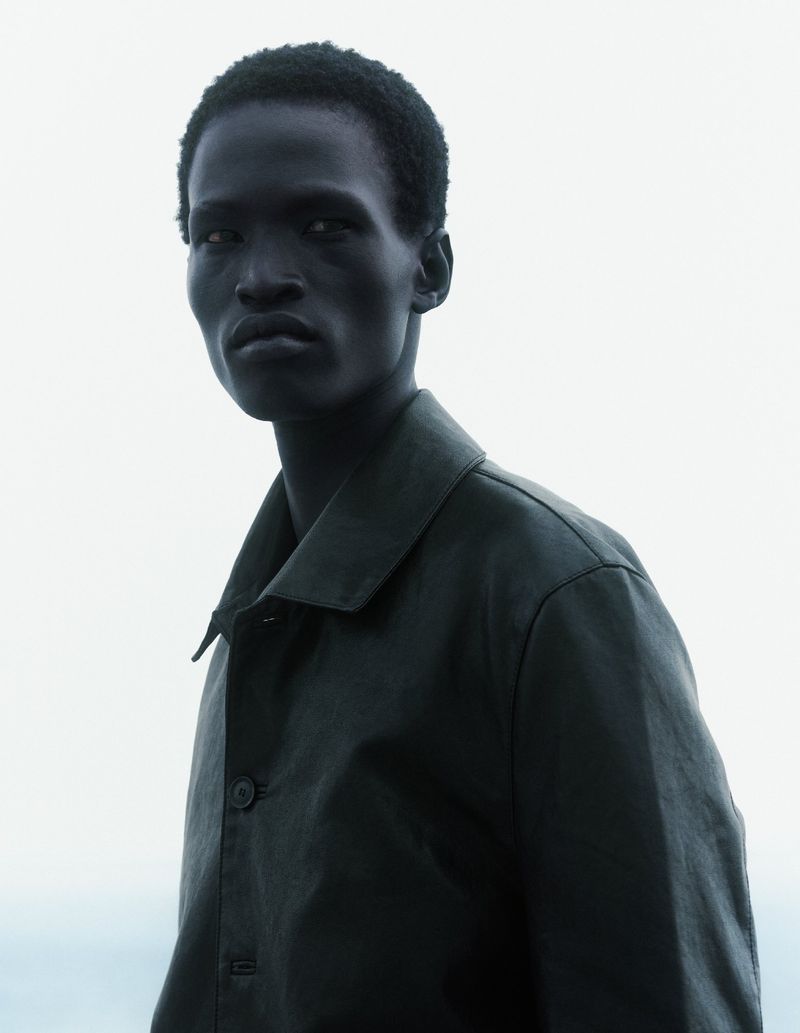 Model Mamuor Majeng wearing a dark collared jacket against a pale overcast sky in a high-contrast fashion portrait.