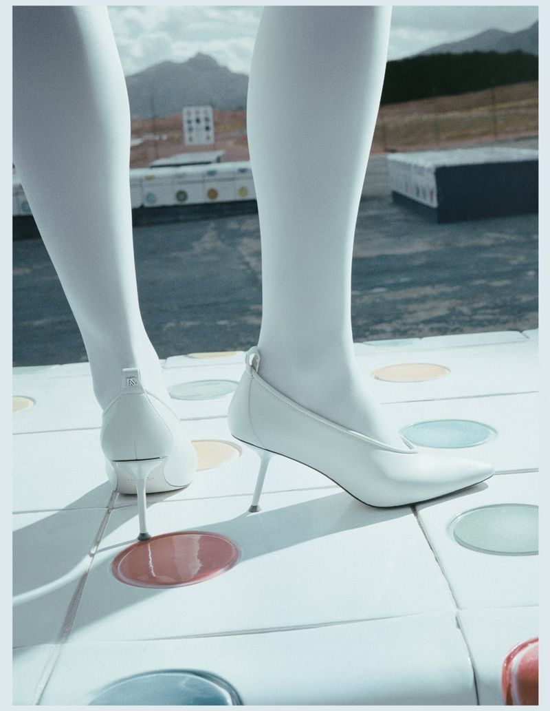 Close-up of model's legs in white tights and white pointed-toe stiletto heels standing on a circular patterned tiled surface outdoors.