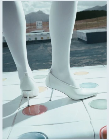 Close-up of model's legs in white tights and white pointed-toe stiletto heels standing on a circular patterned tiled surface outdoors.