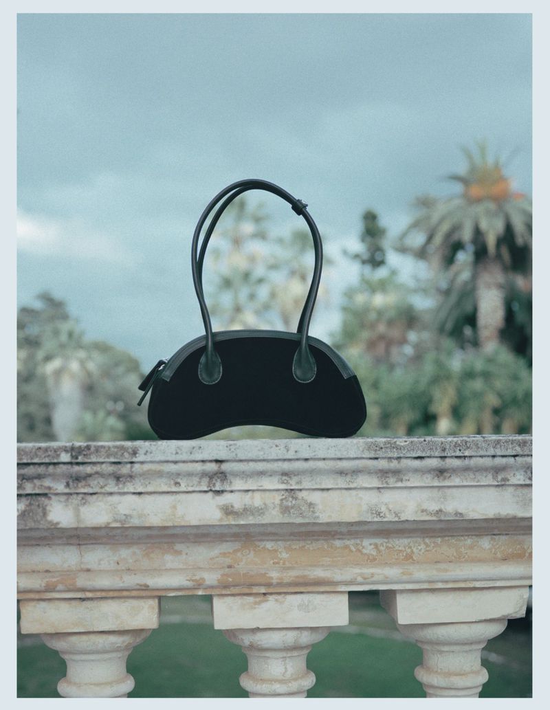 Black handbag with curved shape and top handles on a stone ledge with a blurred background of palm trees and a cloudy sky.