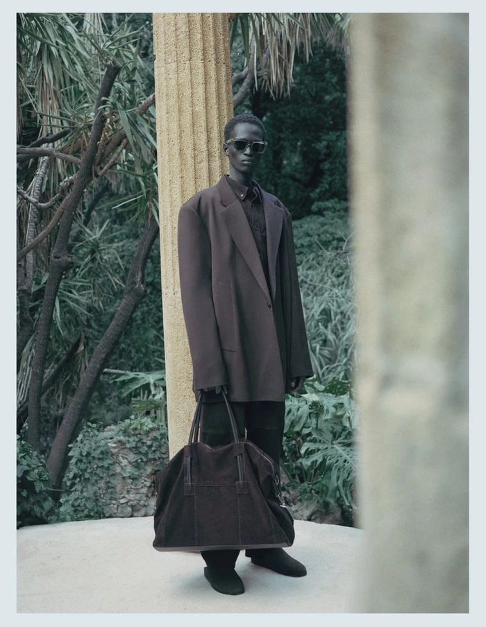 Man wearing a brown oversized blazer and sunglasses holding a large dark duffel bag outdoors among trees.