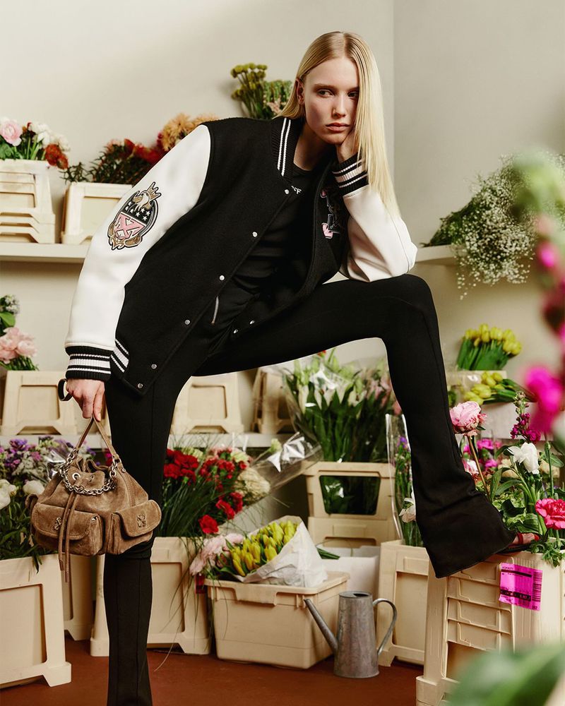 Model Katrine Steiness posing in an Off-White black and white varsity jacket and black pants among crates of fresh flowers.