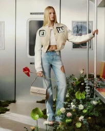 Model Katrine Steiness wearing a cropped varsity jacket and cutout jeans holding a beige handbag inside a subway car filled with plants.