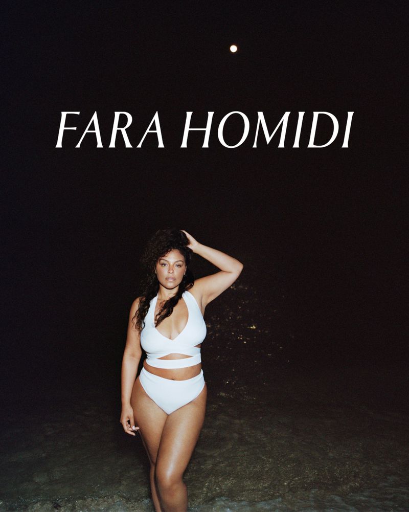 A woman in a white two-piece swimsuit poses on a beach at night under a full moon, with dark, slightly blurred sand and ocean water.