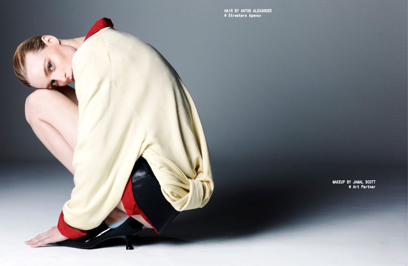 A fashion shot features a woman in a cream jacket and black heels crouching in a studio with a gradient gray background and soft lighting.