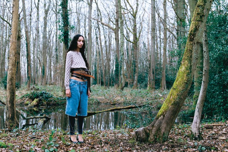 Young woman in cropped sweater, wide-leg jeans, and heels standing in a wooded area with a stream and bare trees in the background.