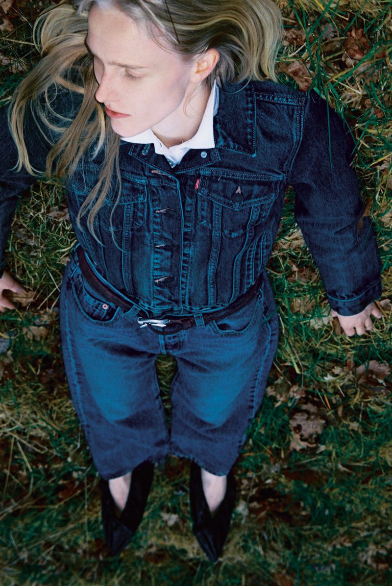 Overhead shot of a young woman with fair skin and long blonde hair lying on grass, wearing a denim jacket and pants, and black shoes.