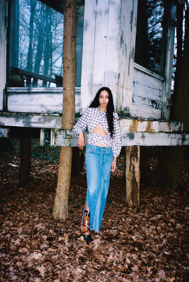 Woman in denim skirt and gingham top poses in a wooded area with an old white structure, possibly a shed, in the background.