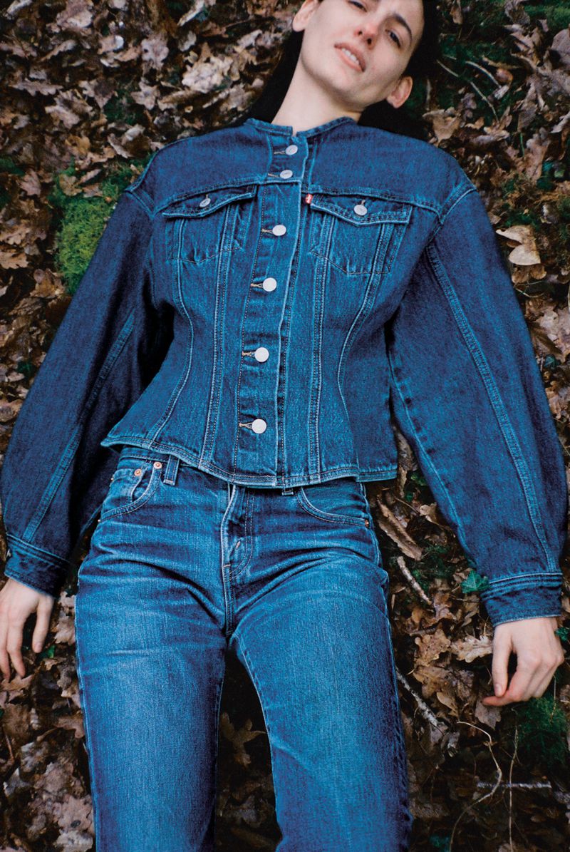 A young adult woman lies on a bed of leaves wearing a blue denim jacket and jeans in an outdoor fashion portrait.