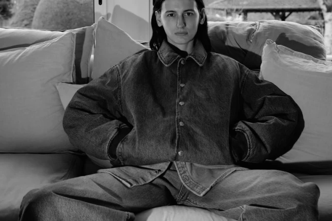 Black and white portrait of an adult female in an oversized denim jacket and pants, sitting on a sofa with throw pillows indoors.