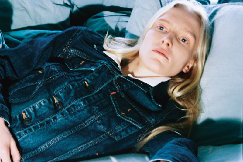 Fair-skinned woman with blonde hair lies on a bed covered with a blue denim jacket in a coolly lit indoor setting.
