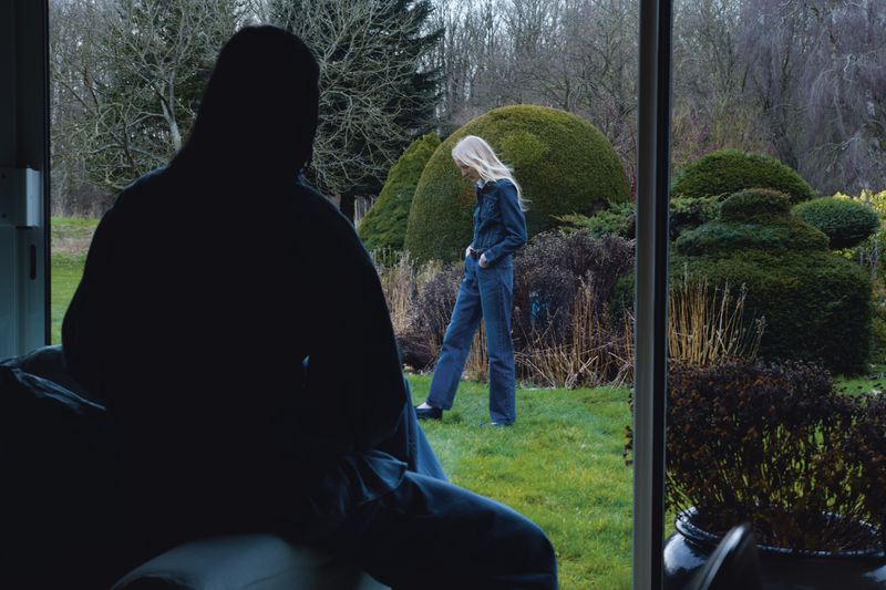 An indoor shot of a woman in a jumpsuit standing in a garden framed by a window, with a silhouetted figure in the foreground.