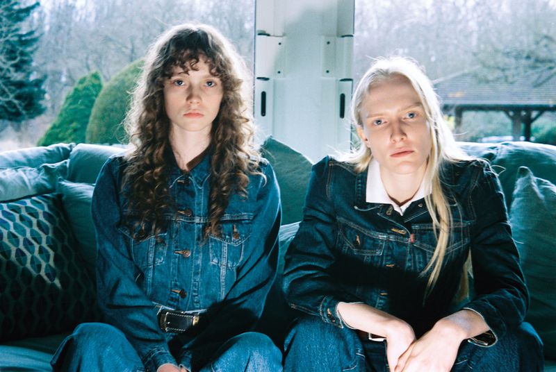 Two young women in matching denim jackets and jeans pose indoors on a teal couch with a window and greenery visible in the background.