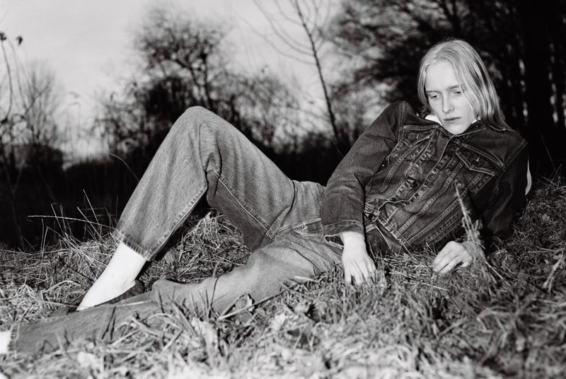 Monochrome image of a reclining woman in denim jacket and jeans on grassy terrain, featuring natural lighting and a somber expression.