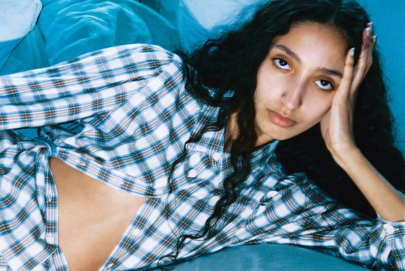 A young woman with long dark curly hair rests her head on her hand while lying on a turquoise surface in a light blue plaid shirt.