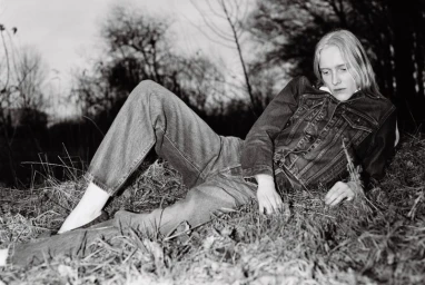 Model wearing a denim jacket and jeans lying in tall grass in a black and white editorial photograph.