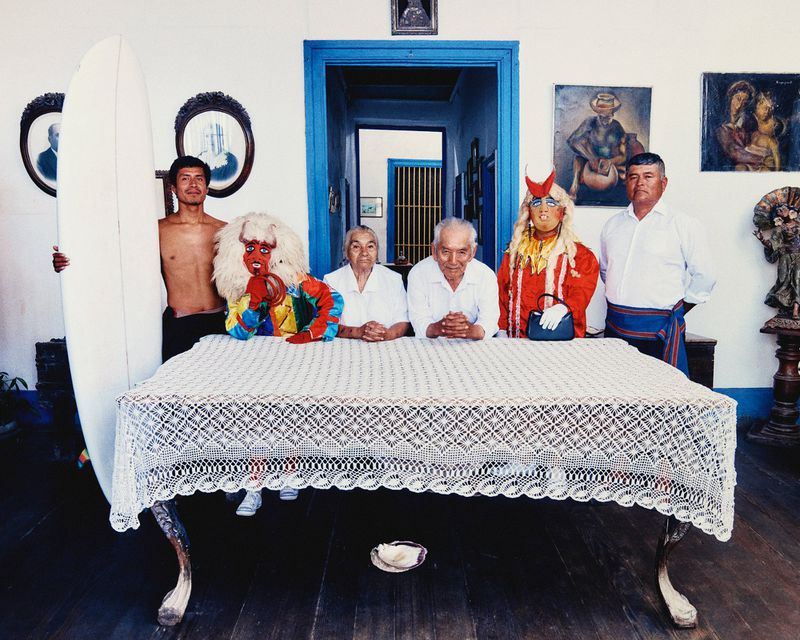Group portrait behind a lace-covered table with an upright surfboard, masked figures, and family members in a traditional home.